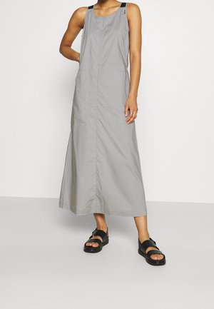 Woman wearing a sleeveless gray maxi dress with black strap details and black open-toe sandals standing against a light background.