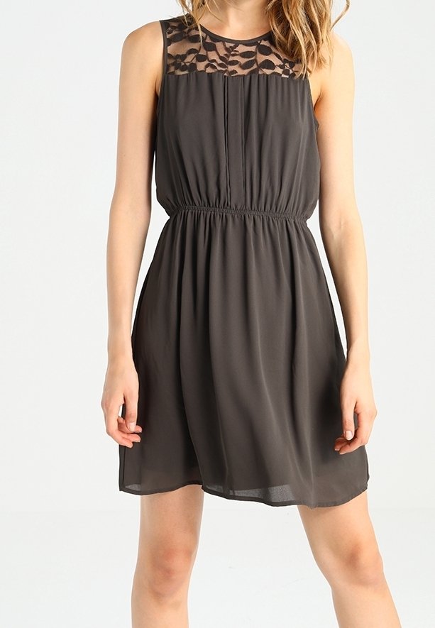 Robe noire sans manches avec empiècement en dentelle florale transparente, taille froncée et jupe fluide longueur genou portée par une personne debout.