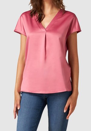 Femme portant un chemisier rose en satin à manches courtes et col en V, associé à un jean bleu en denim, sur un fond clair uni.