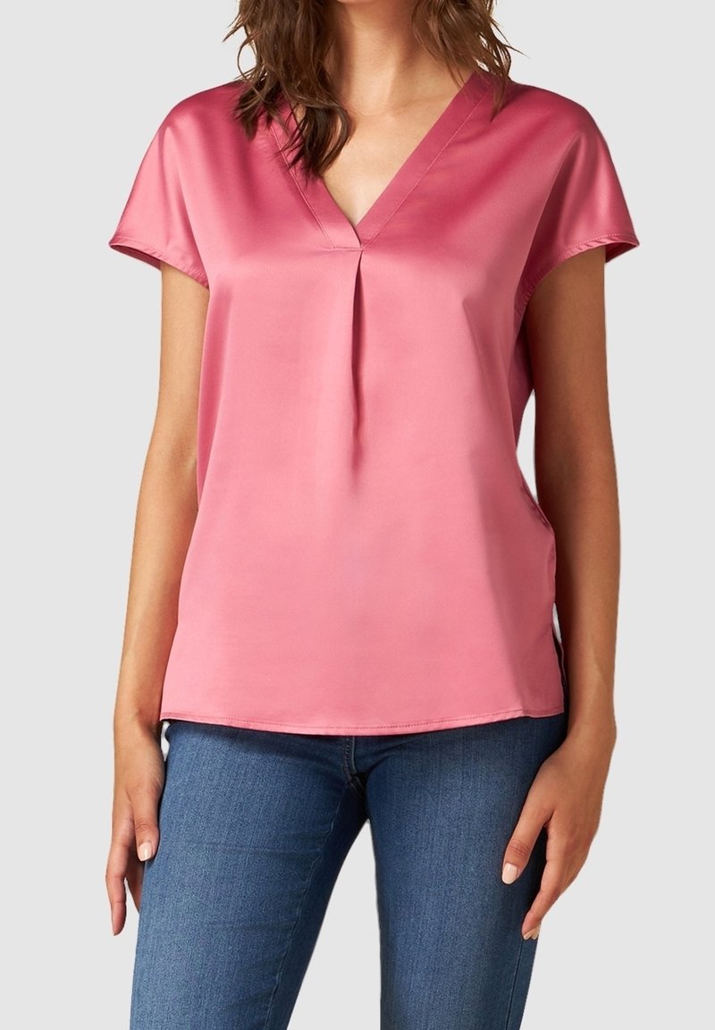 Femme portant un chemisier rose en satin à manches courtes et col en V, associé à un jean bleu en denim, sur un fond clair uni.