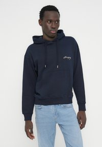 Μπλε-σκούρο φούτερ με κουκούλα από απαλό ύφασμα, με μπροστινή τσέπη, ρυθμιζόμενα κορδόνια και λογότυπο στο στήθος.