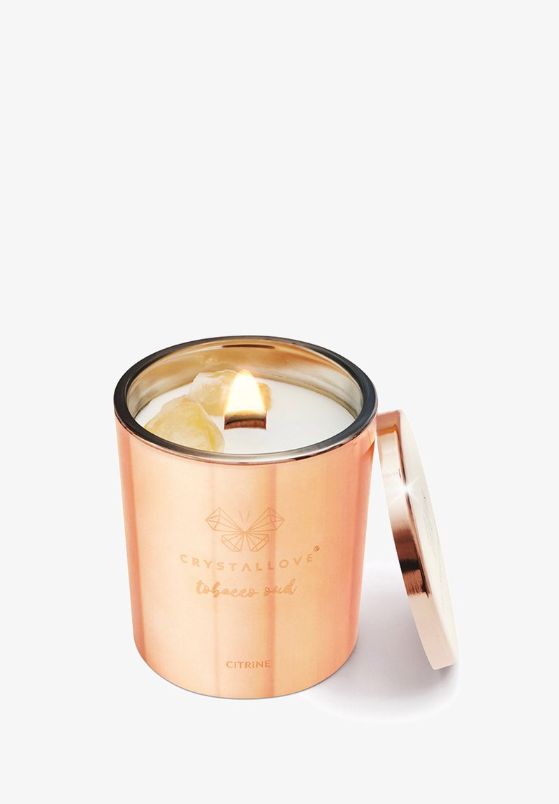 Miedziany pojemnik na świecę z pokrywką, z białym wnętrzem woskowym i migoczącym płomieniem. Zawiera żółte kryształowe akcenty i tłoczone logo.