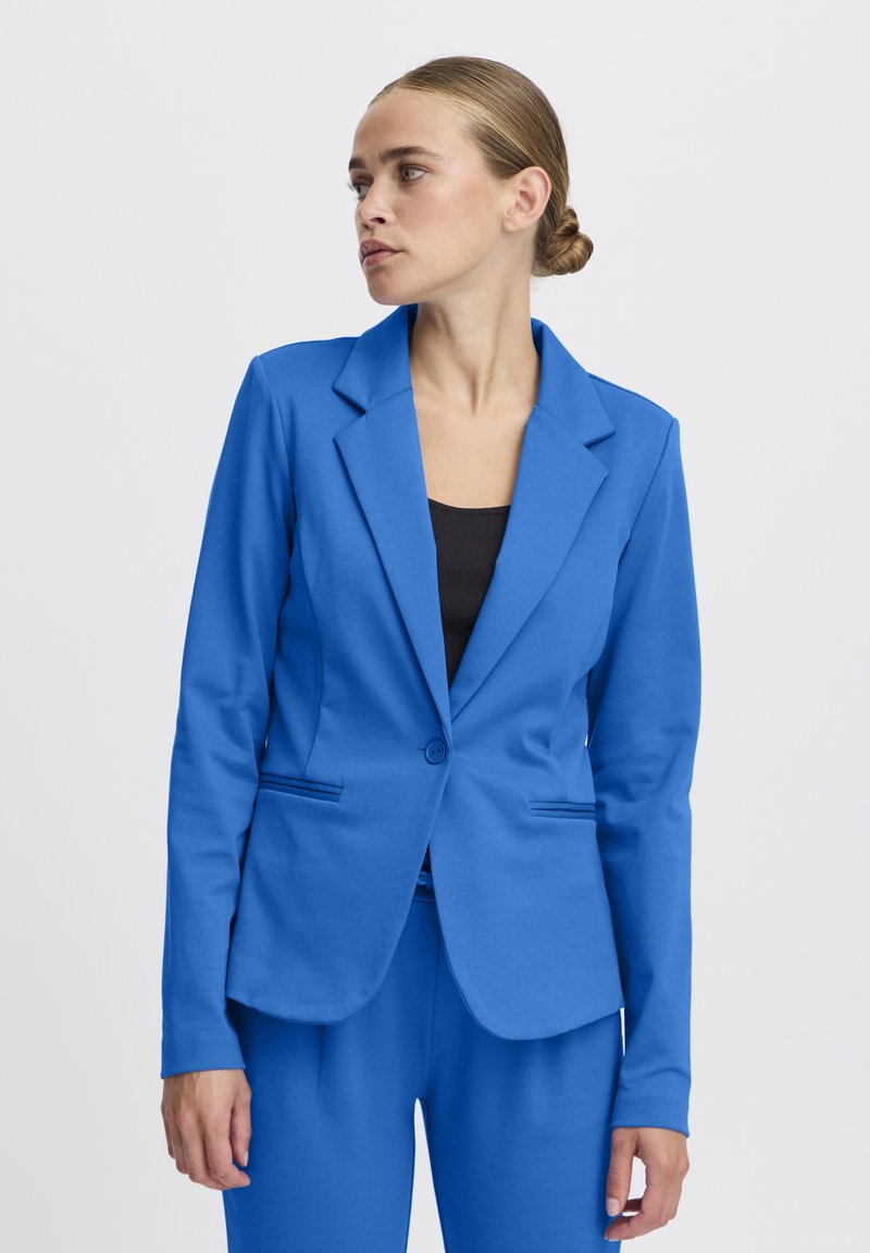 Femme aux cheveux blonds en chignon portant un blazer bleu ajusté et un pantalon assorti, regardant vers la gauche sur un fond uni.