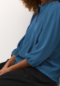 Sininen ribbineulottu paita, jossa korkea kaulus, lyhyet, poimutetut hihat ja rento istuvuus. Kangas on kudottu, pehmeä ja kevyt ulkonäöltään.