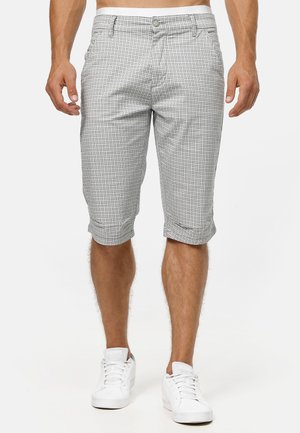 Mandlig model iført grå og hvide ternede knælange shorts og hvide sneakers, stående mod en ensfarvet baggrund.