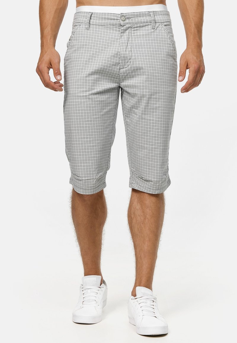 Modèle masculin portant un short à carreaux gris et blanc longueur genoux et des baskets blanches, debout devant un fond uni.