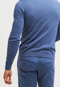 Blauer gestrickter Pullover mit geripptem Saum und Bündchen, kombiniert mit hellblauen Hosen mit geknöpften Gesäßtaschen. Weiche Textur, taillierter Schnitt.