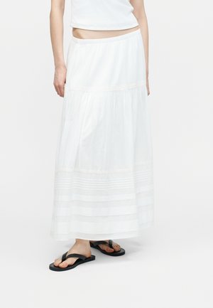 Femme portant une jupe maxi blanche à volants, un débardeur court blanc et des sandales noires à lanières, debout sur un fond uni.