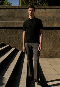 Schwarzes Polohemd mit kurzen Ärmeln, kombiniert mit grauen, gestreiften Hosen und schwarzen Schuhen. Das Modell steht auf Betontreppen vor einer strukturierten Wand.