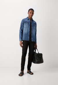 Farmerdzseki zsebbel, fekete, slim-fit farmernadrág, fekete papucs és egy struktúrált fekete kézitáska. Semleges háttér.