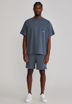 Mężczyzna stojący, ubrany w dopasowaną, ciemnoszarą, oversize'ową koszulkę i szorty oraz białe trampki na jednolitym szarym tle.