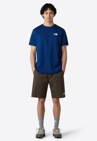 Modré tričko s krátkým rukávem a malým bílým logem, spárované s khaki šortkami a šedými ponožkami. Oblečení doplňují šedé boty.