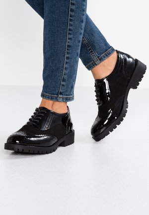 Kiiltävät mustat brogue-kengät paksuilla pohjilla, joita pidetään ylös käännettyjen sinisten farkkujen kanssa valkoisella lattialla, toinen jalka kevyesti kohotettuna.