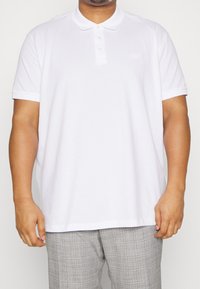 Mężczyzna w gładkiej białej koszulce polo z krótkim rękawem i szarych spodniach w kratę, stojący z luzem, z rękami swobodnie opadającymi wzdłuż ciała.