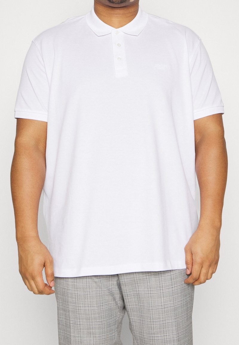 Mężczyzna w gładkiej białej koszulce polo z krótkim rękawem i szarych spodniach w kratę, stojący z luzem, z rękami swobodnie opadającymi wzdłuż ciała.