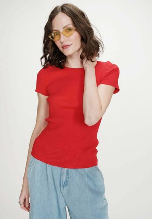 Femme aux cheveux foncés mi-longs portant des lunettes jaunes en forme d'œil de chat, un haut rouge à manches courtes et un jean bleu clair ample, posant devant un fond blanc.