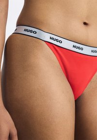 Punainen tanga valkoisella vyötärönauhalla, jossa on "HUGO"-logo. Materiaali näyttää sileältä ja joustavalta, istuen tiiviisti vartaloon.