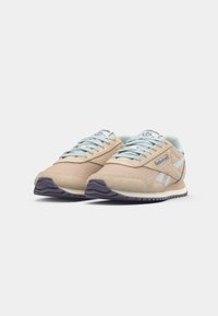 Beigu un baltu Reebok sneakeru pāris ar gaiši zilām šņorēm un tumši pelēkiem zoliem uz baltā fona, skats no leņķa priekšpusē.