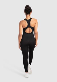 Zwarte eendelige body met een racerback ontwerp en een cirkelvormige uitsnijding op de rug, gemaakt van rekbaar materiaal, gecombineerd met witte sneakers.