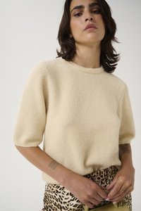Beige, short-sleeve sweater made of soft, textured fabric with a ribbed neckline. Paired with leopard print pants and holding a green bag.
