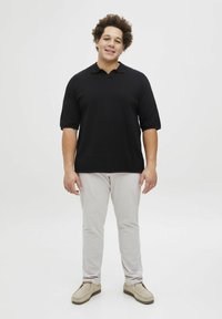 Juodas trumpomis rankovėmis polo marškinėliai su laisvu sijonu, derinami su šviesiai pilkais kelnais ir smėlio spalvos slip-on batais, ant neutralios fono.