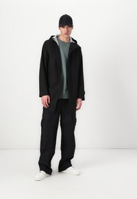 Zwarte waterdichte jas met een grijze capuchon, gedragen over een groene sweatshirt en zwarte cargo broek. Witte sneakers maken de outfit compleet.