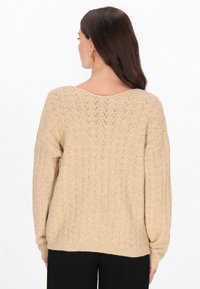 Beżowy sweter dzianinowy o luźnym kroju, z dekoltem w serek, teksturowanym wzorem w romby oraz prążkowanymi mankietami i dolnym brzegiem. Brak dodatkowych informacji.