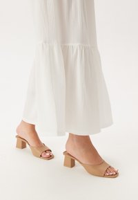 Tan open-toe mules met een vierkante neus en blokhak, gecombineerd met een witte, textuurachtige, vloeiende maxi-jurk met een gerafeld zoom.