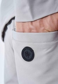 Bolsillo de pantalones de tela gris con una parche circular negro en relieve con "Project X Paris," que presenta una textura suave y costuras sutiles.