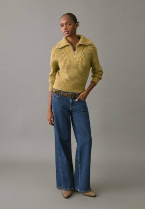 Pull en maille côtelée jaune moutarde avec un col zippé, associé à un jean bleu denim à taille haute et évasé, ainsi qu'à des chaussures à talons beiges.
