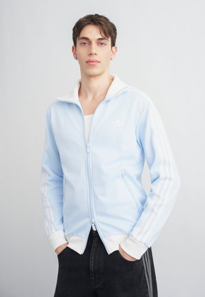 Joven con cabello corto que lleva una chaqueta azul claro con cremallera y rayas blancas en las mangas, de pie con las manos en los bolsillos de los pantalones negros.