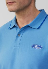 Man in een lichtblauw polo shirt met een donkerblauwe kraag en een Ford-logo geborduurd op de borst.