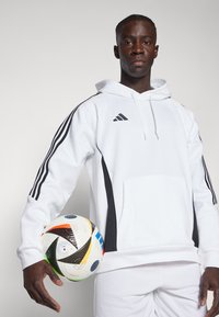 Weißer Hoodie mit schwarzen Seitenelementen und drei schwarzen Streifen an den Ärmeln, mit einer Fronttasche. Hält einen mehrfarbigen Fußball.