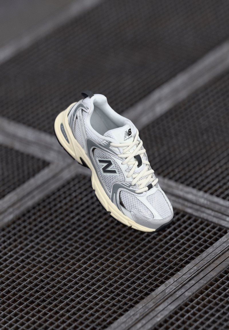 Chaussure de course New Balance blanche et grise avec semelle crème sur une surface en grille métallique sombre.