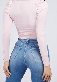 Haut rose à manches longues côtelé avec des épaules bouffantes, associé à un jean en denim bleu clair avec des poches arrière et des détails de boutons sur les manches.