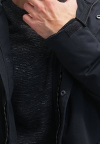 Veste zippée noire avec un pull en maille gris texturé en dessous. La veste présente des boutons-pression et des poignets doux ; le pull a un design subtil en couches.