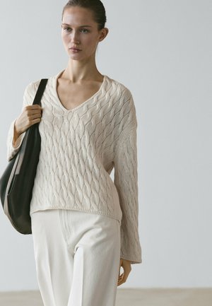 Žena v krémovém svetr s výstřihem do V a bílých kalhotách, nesoucí velkou černou kabelku přes rameno na jednobarevném světle pozadí.