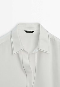 Camicia bianca con colletto classico, dettagli di cucitura nera lungo il colletto e la patta, realizzata in un tessuto liscio.