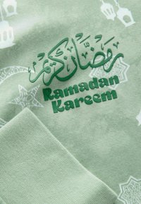 Green fabric featuring "Ramadan Kareem" in raised green lettering. Background includes white geometric patterns and lantern designs.