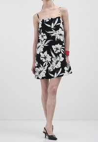 Žena v černobílých květinových šatech bez rukávů s tenkými ramínky, černých sandálech na podpatku a červeném náramku.