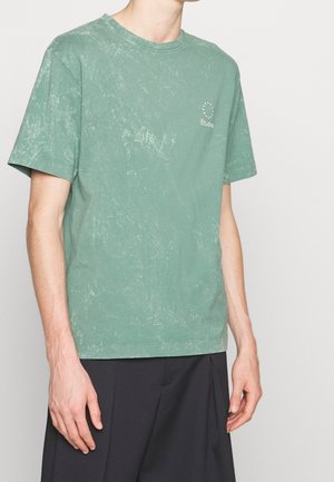 Osoba ubrana w wyblakły zielony T-shirt z krótkim rękawem i małym białym logo "Etudes" na klatce piersiowej, w połączeniu z luźnymi czarnymi spodniami.