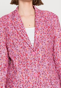 Blazer rose à motifs avec des dessins floraux, des boutons roses, un col à revers cranté et un tissu texturé.