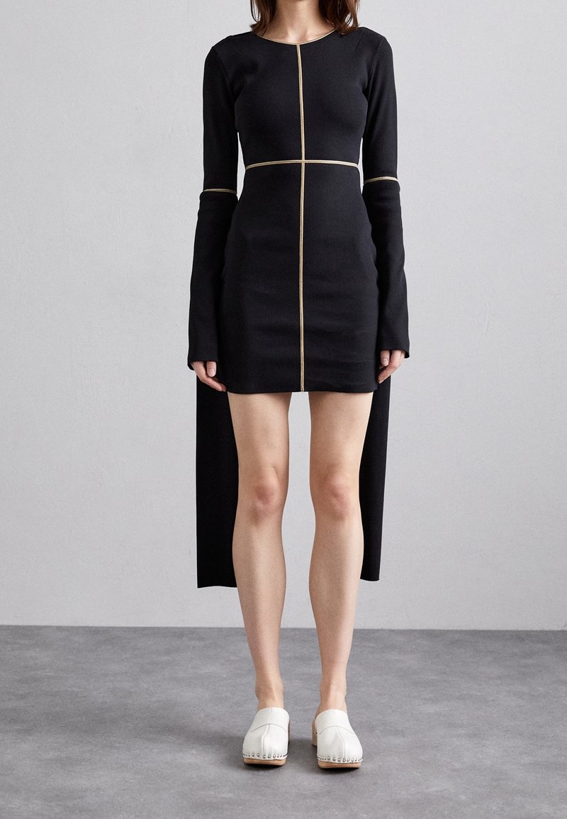 Svart långärmad klänning med en figursydd silhuett, guldtråd och mittlång splitt bak. Matchas med vita träskor.