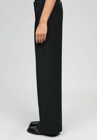 Personne portant un pantalon noir taille haute à jambes larges et des mocassins brillants noirs, se tenant de profil sur un fond clair uni.