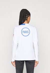 T-shirt de sport à manches longues, blanc, avec un accent bleu au niveau de l'encolure et un grand texte circulaire "PARIS" dans le dos.