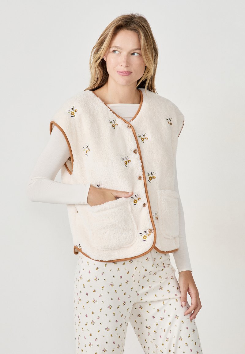 Gilet in pelliccia sintetica color crema con bordi marroni e ricami floreali. Presenta una apertura sul davanti, maniche a cappuccio e due tasche frontali.