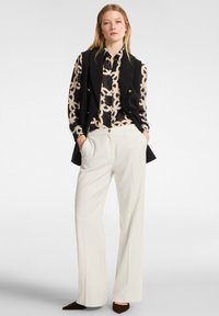 Blazer negro, blusa beige con un patrón, pantalones blancos. El blazer tiene botones dobles y bolsillos; la blusa presenta un diseño geométrico. Planas marrones.