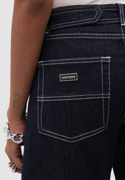 Poche de jean bleu foncé avec surpiqûres blanches, une petite étiquette rectangulaire "WRSTBHVR", et une main portant des bijoux argentés épais à proximité.