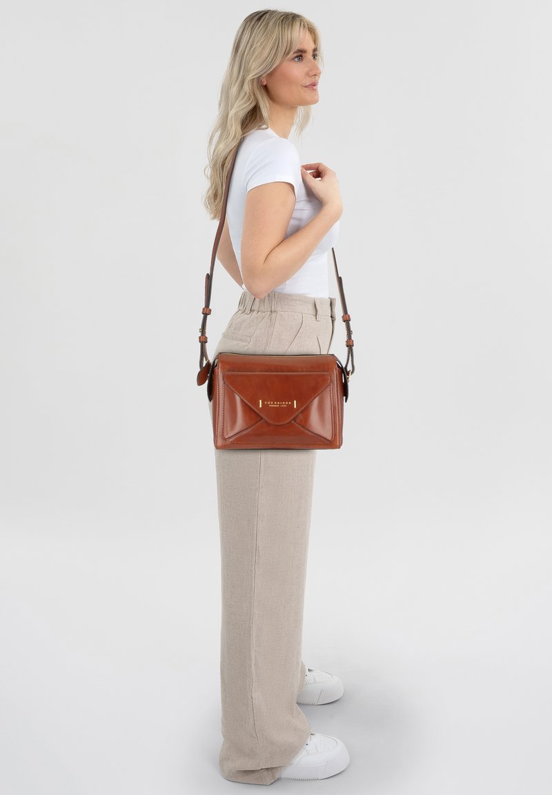 Braune Leder-Umhängetasche mit strukturiertem Design, goldenen Akzenten und einem umklappbaren Deckel. Das Modell trägt beigefarbene Hose und weiße Sneakers.