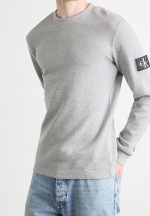 Mężczyzna w jasnoszarej koszuli z długim rękawem z naszywką logo Calvin Klein na rękawie i jasnoniebieskich dżinsach z ręką w kieszeni.